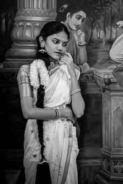 A woman in traditional attire stands pensively in front of a painting. She is adorned with jewelry and a long braid decorated with flowers. The background features a classical-style painted image of another woman in a similar pose.