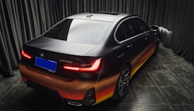 A luxury sedan with a sleek design and matte finish, parked indoors. The car features a unique gradient paint job transitioning from black to red and yellow towards the bottom. The taillights are illuminated, creating a striking contrast with the dark surroundings. The floor is made of glossy black tiles, and the background consists of dark curtains.