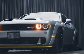 A sleek gray sports car with prominent front-facing features such as a bold grille and illuminated headlights. The car is positioned in front of a dark curtain backdrop, emphasizing its shiny exterior and aggressive design. The visible badge indicates a high-performance model.