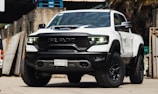 A white pickup truck with a rugged design and large tires is parked in an industrial setting. The vehicle features prominent RAM branding on the front grille and has a hood scoop. The truck's headlights are illuminated, casting a bright light. The surroundings include concrete walls and stacked wooden pallets.