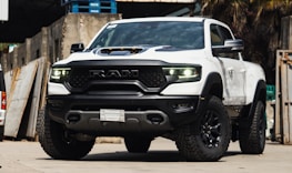 A white pickup truck with a rugged design and large tires is parked in an industrial setting. The vehicle features prominent RAM branding on the front grille and has a hood scoop. The truck's headlights are illuminated, casting a bright light. The surroundings include concrete walls and stacked wooden pallets.