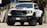A white pickup truck with a rugged design and large tires is parked in an industrial setting. The vehicle features prominent RAM branding on the front grille and has a hood scoop. The truck's headlights are illuminated, casting a bright light. The surroundings include concrete walls and stacked wooden pallets.