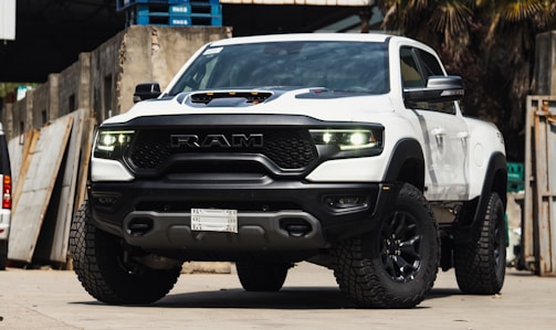 A white pickup truck with a rugged design and large tires is parked in an industrial setting. The vehicle features prominent RAM branding on the front grille and has a hood scoop. The truck's headlights are illuminated, casting a bright light. The surroundings include concrete walls and stacked wooden pallets.