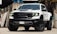 A white pickup truck with a rugged design and large tires is parked in an industrial setting. The vehicle features prominent RAM branding on the front grille and has a hood scoop. The truck's headlights are illuminated, casting a bright light. The surroundings include concrete walls and stacked wooden pallets.