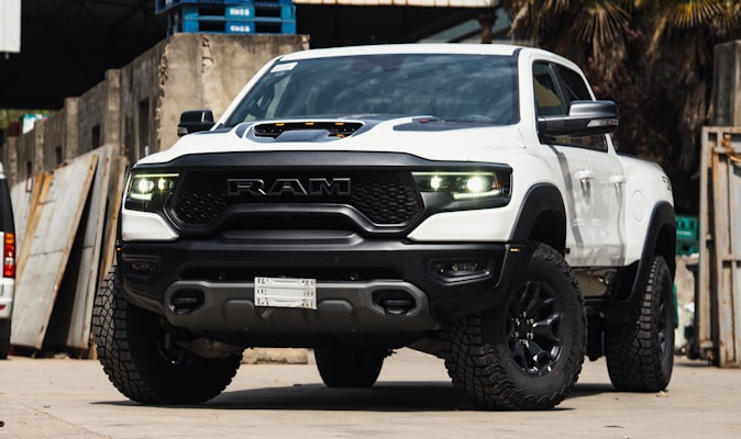 A white pickup truck with a rugged design and large tires is parked in an industrial setting. The vehicle features prominent RAM branding on the front grille and has a hood scoop. The truck's headlights are illuminated, casting a bright light. The surroundings include concrete walls and stacked wooden pallets.