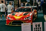 A red racing car with prominent branding and sponsors is displayed at an indoor event. The car is surrounded by people who appear to be attendees, some of whom are looking at the car and walking around. Various items and promotional materials are visible in the foreground, indicating a possible exhibition or show.