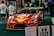 A red racing car with prominent branding and sponsors is displayed at an indoor event. The car is surrounded by people who appear to be attendees, some of whom are looking at the car and walking around. Various items and promotional materials are visible in the foreground, indicating a possible exhibition or show.