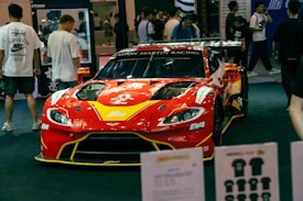 A red racing car with prominent branding and sponsors is displayed at an indoor event. The car is surrounded by people who appear to be attendees, some of whom are looking at the car and walking around. Various items and promotional materials are visible in the foreground, indicating a possible exhibition or show.