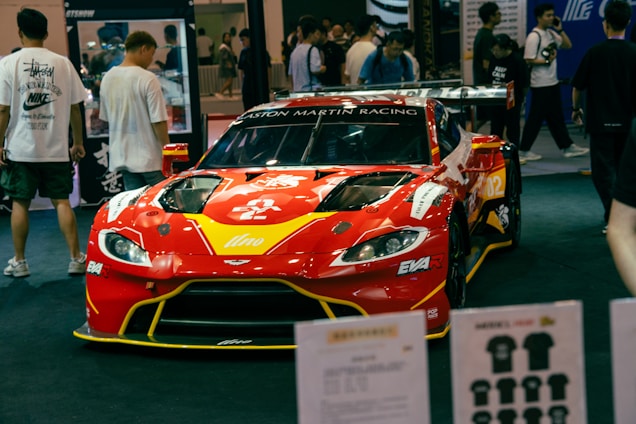 A red racing car with prominent branding and sponsors is displayed at an indoor event. The car is surrounded by people who appear to be attendees, some of whom are looking at the car and walking around. Various items and promotional materials are visible in the foreground, indicating a possible exhibition or show.