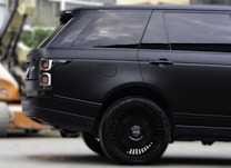 A sleek black SUV with tinted windows, featuring a modern and luxurious design. The focus is on the rear side and the distinctive alloy wheels, while the background is slightly blurred, indicating an urban setting.