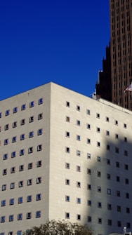 A modern building with a white facade displaying rows of small, square windows. The windows have red borders, and the building casts a shadow on an adjacent structure. A deep blue sky forms the background, and there is an American flag visible at the top right corner.