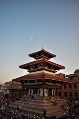 A traditional tiered temple with intricate wooden carvings is surrounded by people sitting on its steps. Motorbikes are parked in front of the temple, and a busy crowd walks by. The evening sky is clear with a visible half moon, and the atmosphere is vibrant with activity.