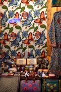 A richly decorated scene featuring a tapestry adorned with colorful floral and butterfly patterns in vibrant shades of orange, blue, and green. In front, an assortment of intricately painted ceramic items, including vases, bowls, and boxes, are displayed. A patterned bag with pink embroidery is prominently positioned at the bottom. Adjacent is a garment with floral motifs, hinting at artisanal craftsmanship.
