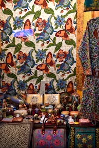 A richly decorated scene featuring a tapestry adorned with colorful floral and butterfly patterns in vibrant shades of orange, blue, and green. In front, an assortment of intricately painted ceramic items, including vases, bowls, and boxes, are displayed. A patterned bag with pink embroidery is prominently positioned at the bottom. Adjacent is a garment with floral motifs, hinting at artisanal craftsmanship.