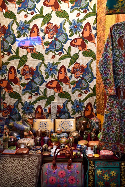 A richly decorated scene featuring a tapestry adorned with colorful floral and butterfly patterns in vibrant shades of orange, blue, and green. In front, an assortment of intricately painted ceramic items, including vases, bowls, and boxes, are displayed. A patterned bag with pink embroidery is prominently positioned at the bottom. Adjacent is a garment with floral motifs, hinting at artisanal craftsmanship.