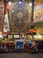 A vibrant display of handmade crafts and textiles including rugs, pillows, and wooden carvings. The items exhibit a variety of colors and patterns, with motifs depicting animals like cats and elephants. Several brightly lit light bulbs hang overhead, illuminating the scene. Payment signs for Visa and Mastercard are visible, suggesting items for sale.