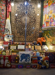 A vibrant display of handmade crafts and textiles including rugs, pillows, and wooden carvings. The items exhibit a variety of colors and patterns, with motifs depicting animals like cats and elephants. Several brightly lit light bulbs hang overhead, illuminating the scene. Payment signs for Visa and Mastercard are visible, suggesting items for sale.
