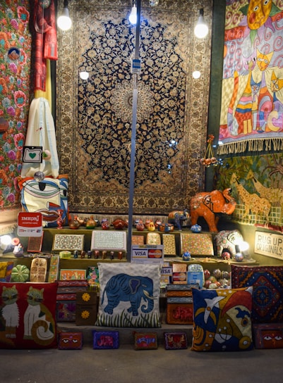 A vibrant display of handmade crafts and textiles including rugs, pillows, and wooden carvings. The items exhibit a variety of colors and patterns, with motifs depicting animals like cats and elephants. Several brightly lit light bulbs hang overhead, illuminating the scene. Payment signs for Visa and Mastercard are visible, suggesting items for sale.
