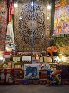 A vibrant display of handmade crafts and textiles including rugs, pillows, and wooden carvings. The items exhibit a variety of colors and patterns, with motifs depicting animals like cats and elephants. Several brightly lit light bulbs hang overhead, illuminating the scene. Payment signs for Visa and Mastercard are visible, suggesting items for sale.