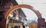 An archway with text written in a South Asian script, leading into a temple complex surrounded by traditional brick buildings. People can be seen at the edge of the frame. The sun is shining brightly, casting lens flares.