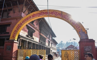 An archway with text written in a South Asian script, leading into a temple complex surrounded by traditional brick buildings. People can be seen at the edge of the frame. The sun is shining brightly, casting lens flares.