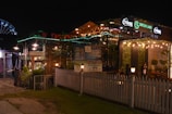 A nighttime scene of a vibrant outdoor restaurant or cafe with lights adorning the area. The establishment has signage for 'Green Lake' and 'Tuborg', and is surrounded by white picket fencing and greenery. There are people visible, suggesting a lively atmosphere.