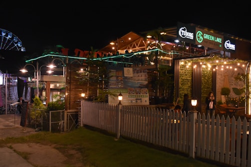 A nighttime scene of a vibrant outdoor restaurant or cafe with lights adorning the area. The establishment has signage for 'Green Lake' and 'Tuborg', and is surrounded by white picket fencing and greenery. There are people visible, suggesting a lively atmosphere.