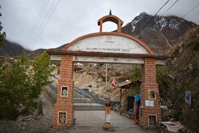 A rustic entrance gate constructed with reddish-brown bricks leads to a mountainous area. The archway is adorned with various symbols and inscriptions, including a welcoming message to the Muktinath Temple. Surrounding the gate are rocky terrain and sparse vegetation, with mist-covered mountains looming in the background.