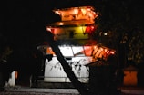 A traditional multi-tiered structure is illuminated by string lights at night, casting a warm glow. The building is surrounded by trees, with shadows creating a mysterious atmosphere.