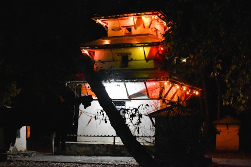A traditional multi-tiered structure is illuminated by string lights at night, casting a warm glow. The building is surrounded by trees, with shadows creating a mysterious atmosphere.