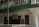 A two-story building with a green and white exterior features a small business sign that reads 'Ms Tashi Monpi Store, Urgelling, Tawang.' The upper level has a decorative railing with green accents, and the ground floor appears to have closed wooden doors and windows.