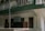 A two-story building with a green and white exterior features a small business sign that reads 'Ms Tashi Monpi Store, Urgelling, Tawang.' The upper level has a decorative railing with green accents, and the ground floor appears to have closed wooden doors and windows.