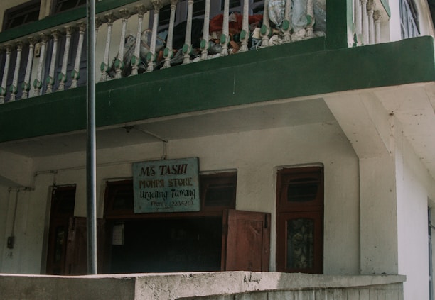A two-story building with a green and white exterior features a small business sign that reads 'Ms Tashi Monpi Store, Urgelling, Tawang.' The upper level has a decorative railing with green accents, and the ground floor appears to have closed wooden doors and windows.