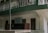 A two-story building with a green and white exterior features a small business sign that reads 'Ms Tashi Monpi Store, Urgelling, Tawang.' The upper level has a decorative railing with green accents, and the ground floor appears to have closed wooden doors and windows.