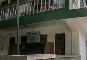 A two-story building with a green and white exterior features a small business sign that reads 'Ms Tashi Monpi Store, Urgelling, Tawang.' The upper level has a decorative railing with green accents, and the ground floor appears to have closed wooden doors and windows.