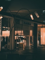 Dimly lit storefront of a clothing shop with the brand name visible above the entrance. The closed shop is surrounded by dark and subdued lighting, with some illuminated fixtures creating a warm glow. Metal structures or decor elements are placed in the foreground.