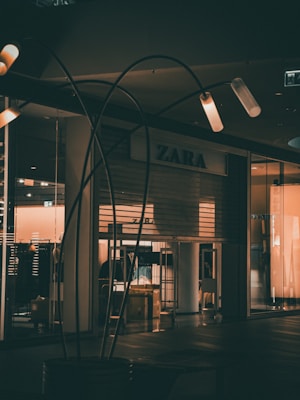 Dimly lit storefront of a clothing shop with the brand name visible above the entrance. The closed shop is surrounded by dark and subdued lighting, with some illuminated fixtures creating a warm glow. Metal structures or decor elements are placed in the foreground.