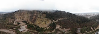Aerial view of a large quarry surrounded by dense forest. The quarry has visible layers of rock in the cliffs and is partially covered by low-hanging clouds. Roads and structures are visible at the base of the quarry. The surrounding area is heavily forested, with a view extending to a distant city skyline on the right.