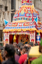 An intricately decorated chariot with vibrant and detailed designs is paraded through a street. The chariot features colorful patterns, religious motifs, and is topped with a dome-like structure. A statue or idol is visible inside the chariot, adorned with garlands. A crowd of people surrounds the chariot, suggesting a festive or religious event.
