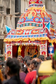 An intricately decorated chariot with vibrant and detailed designs is paraded through a street. The chariot features colorful patterns, religious motifs, and is topped with a dome-like structure. A statue or idol is visible inside the chariot, adorned with garlands. A crowd of people surrounds the chariot, suggesting a festive or religious event.