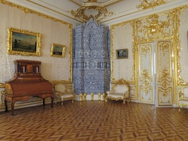 A luxurious room featuring ornate decorations, with gilded walls and intricate patterns. There are classic-style furniture pieces including a wooden roll-top desk and elegant upholstered chairs. The blue and white tiled stove is a focal point, surrounded by richly detailed gold accents.