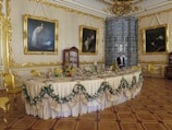 An opulent dining room with an elegantly set table adorned with draped fabric and floral garlands. The walls are decorated with gold embellishments and large framed paintings. A cabinet holds fine china, and a mannequin dressed in formal attire stands in front of a large decorative fireplace.