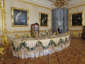 An opulent dining room with an elegantly set table adorned with draped fabric and floral garlands. The walls are decorated with gold embellishments and large framed paintings. A cabinet holds fine china, and a mannequin dressed in formal attire stands in front of a large decorative fireplace.
