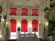 A luxurious, ornately decorated interior with rich red curtains draping down from large windows. The intricate white moldings and carvings adorn the walls and ceiling, creating an elegant and opulent atmosphere. A grand staircase lined with decorative vases and lighting leads to the upper level. Abundant floral arrangements add a touch of nature to the sophisticated setting.