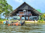 A modern, open-structure architecture with a triangular roof sits at the edge of a body of water. The building has large glass windows and is surrounded by lush greenery, with trees on one side and a small boat anchored near a platform extending into the water.