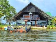 A modern, open-structure architecture with a triangular roof sits at the edge of a body of water. The building has large glass windows and is surrounded by lush greenery, with trees on one side and a small boat anchored near a platform extending into the water.