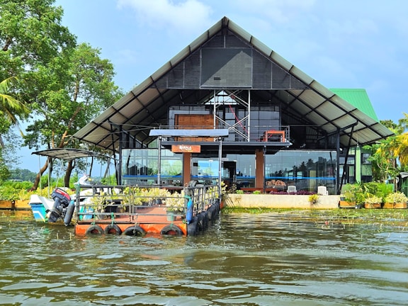 A modern, open-structure architecture with a triangular roof sits at the edge of a body of water. The building has large glass windows and is surrounded by lush greenery, with trees on one side and a small boat anchored near a platform extending into the water.
