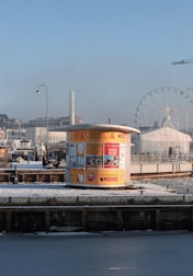 A small yellow kiosk stands on a snowy waterfront, advertising sightseeing cruises and bus tours. The background includes a large Ferris wheel, city buildings, and a construction crane. A person walks by the kiosk, which contrasts against the clear blue sky.