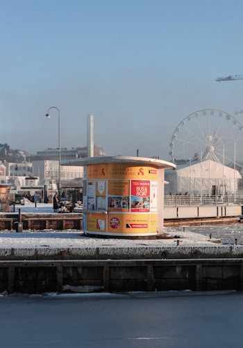 A small yellow kiosk stands on a snowy waterfront, advertising sightseeing cruises and bus tours. The background includes a large Ferris wheel, city buildings, and a construction crane. A person walks by the kiosk, which contrasts against the clear blue sky.