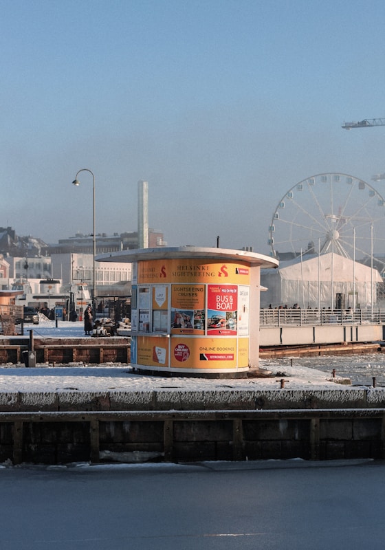 A small yellow kiosk stands on a snowy waterfront, advertising sightseeing cruises and bus tours. The background includes a large Ferris wheel, city buildings, and a construction crane. A person walks by the kiosk, which contrasts against the clear blue sky.